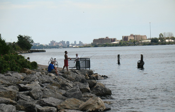 Fishermen at Barretto Point Park in the Bronx, with the buildings of Rikers Island in the distance. A sewage plant, floating correctional facility and half-dozen waste transfer stations are nearby, and the area hosts heavy truck traffic.